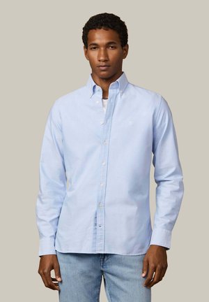 Hombre con una camisa azul clara abotonada sobre una camiseta blanca y jeans azules, de pie frente a un fondo beige liso.