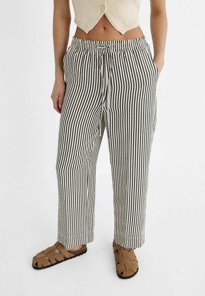 Persoon draagt zwart-witte verticale gestreepte broek met trekkoord aan de taille, crème cropped top met knopen en bruine gevlochten sandalen.