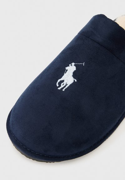 Jūras zila čība ar mīkstu zamša materiālu, noapaļota forma un balta izšūta logo ar jātnieku figūru. Redzama šuvju līnija pa malu.