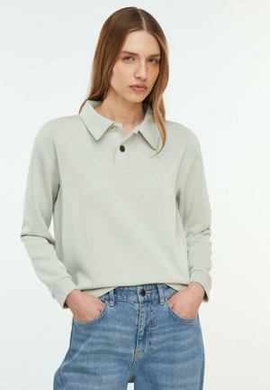 Kobieta z prostymi jasnobrązowymi włosami, ubrana w jasnoszarą, długorękawową koszulę z kołnierzykiem oraz niebieskie dżinsy, ręce w kieszeniach, neutralne tło.