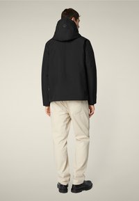 Hombre vestido con una chaqueta con capucha negra, pantalones beige y botas negras, de pie de espaldas contra un fondo neutro.