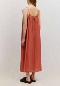 Maxikleid aus weichem rostfarbenem Stoff mit plissierter Textur, verstellbaren Spaghetti-Trägern und einer Bindeschließe auf dem Rücken. Tragen mit schwarzen Sandalen.