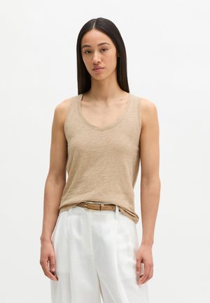 Frau mit glatten dunklen Haaren, die ein beiges ärmelloses Oberteil trägt, das in weiße hoch taillierte Hosen mit einem braunen Gürtel gesteckt ist.