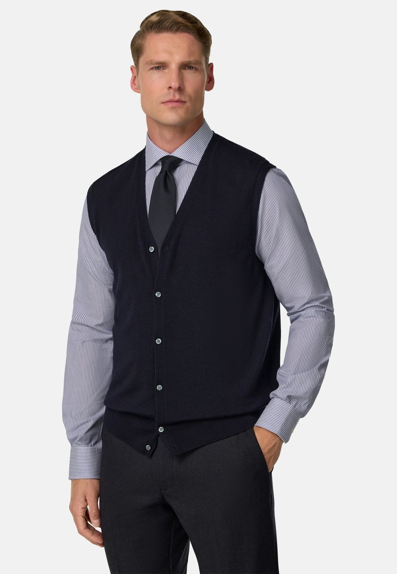 Námořnická pletená vesta s výstřihem do V, s knoflíkovým zapínáním, nošená přes světle modrou pruhovanou košili a doplněná o tmavé kalhoty.