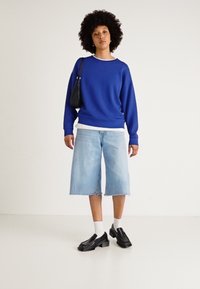 Felpa blu, bordo bianco, pantaloni di jeans blu chiaro, mocassini neri e calzini bianchi. Sfondo semplice. Vestibilità semplice e rilassata.