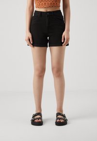 Shorts de mezclilla negra con dobladillo doblado, diseño de cinco bolsillos y cintura alta, combinados con sandalias negras que tienen tachuelas plateadas.