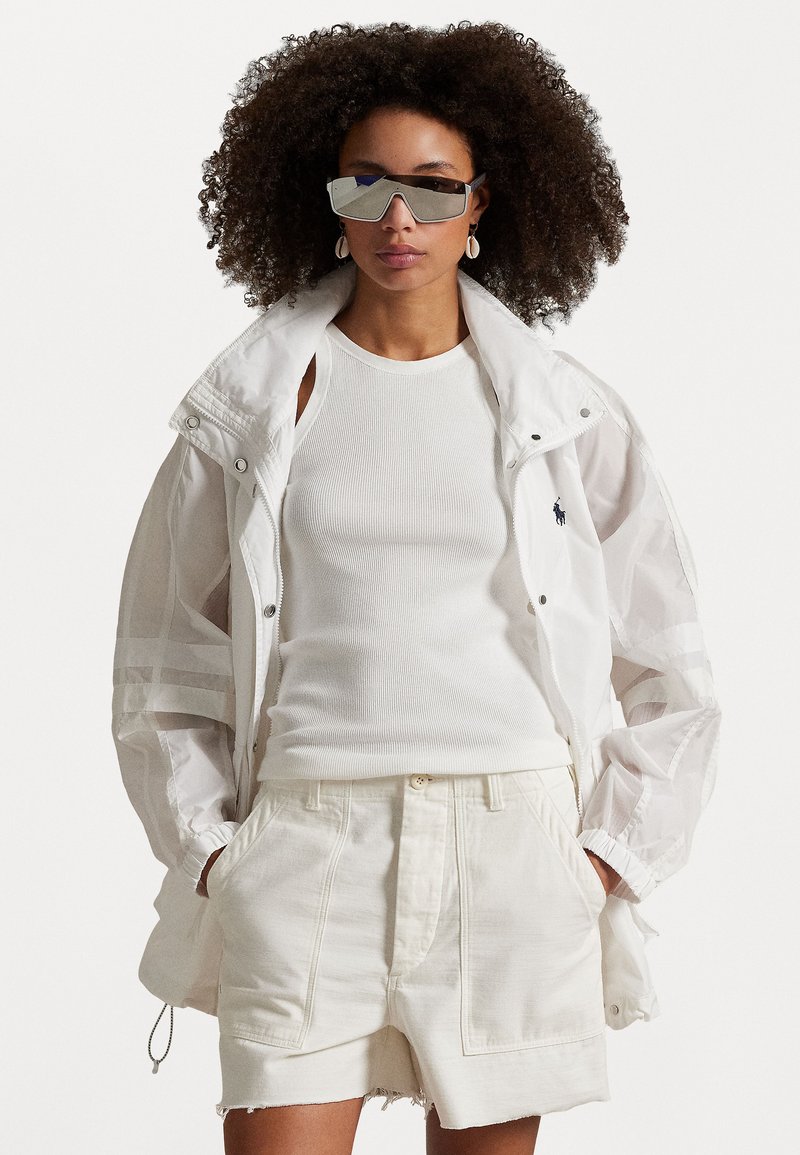 Weiße Lichtsjacke mit reflektierenden Akzenten, weißes geripptes Tanktop, weiße Shorts mit Taschen und verspiegelte Sonnenbrille. Lockige Haare sichtbar.