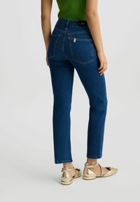 Jeans en denim bleu avec une coupe ajustée, deux poches arrière et un détail de fermeture éclair. Associés à des chaussures plates dorées. Fond uni.