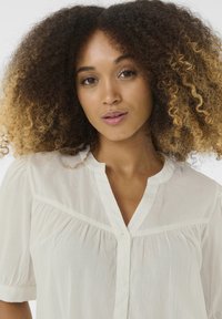 Blusa leggera e trasparente in morbido tessuto bianco, con chiusura a bottoni sul davanti, vestibilità rilassata e maniche corte a sbuffo con delicate arricciature.