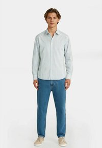 Ung mann står iført en hvit skjorte med lange ermer og tynne vertikale striper, blå jeans og beige sneakers på en hvit bakgrunn.