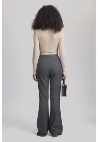 Persoon met krullend haar staat met de rug naar ons toe, draagt een doorschijnend lange mouwen top, hooggerande grijze broek en houdt een kleine handtas vast.