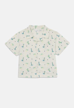 Camisa de botão de meia manga em tecido bege claro com um padrão de dinossauros e palmeiras em verde e azul. Possui colarinho clássico e botões frontais.