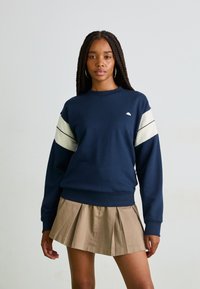 Sweatshirt bleu marine avec des rayures diagonales crème sur les manches, encolure ras du cou, poignets et ourlet côtelés, associé à une jupe plissée beige.