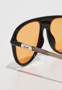 Svarte solbriller med oransje linser, med en kraftig design og transparente armer. "QUAY"-logoen er preget på metallakselen.