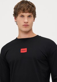 Camiseta negra de manga larga con un parche rectangular rojo que lleva el texto "HUGO" en blanco. Confeccionada en un tejido suave, cuello redondo y corte relajado.