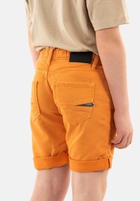 Shorts di jeans arancioni con risvolti, dotati di due tasche posteriori e un dettaglio con zip sulla tasca destra. Dettaglio della cintura nera.