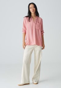 Camicia a maniche lunghe rosa chiaro con colletto a risvolto e tasca frontale, abbinata a pantaloni larghi cremosi dalla texture e scarpe bianche slip-on.