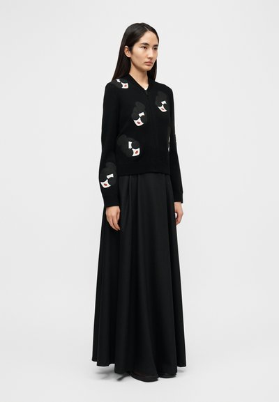 Crni kardigan na patentni zatvarač, s izvezenim grafičkim uzorcima. Uparen s dugom crnom suknjom koja ima glatku teksturu i lagani pad.