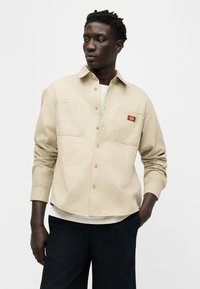 Homme portant une chemise à rayures beige boutonnée par-dessus un t-shirt blanc et un pantalon noir, debout avec une main dans la poche, regardant vers la gauche.