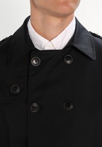 Svart dubbelknäppt kappa i slät tyg, med stora svarta knappar och en bred krage, lagd över en vit skjorta.