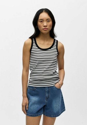 Jeune femme debout portant un débardeur rayé noir et blanc et un short en jean bleu, une main dans la poche, expression neutre.