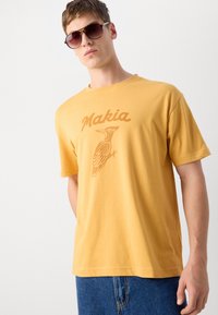 Gul bomull T-skjorte med avslappet passform, med en brun grafikk av en fugl og ordet "Makia" trykt over. Brukt med denimshorts.