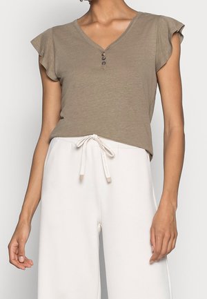 Frau trägt ein taubenblaues kurzärmeliges V-Ausschnitt-Oberteil mit drei Knöpfen und weiße hochtaillierte Hose mit Kordelzug.