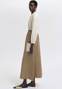 Lang, beige kjole med en afslappet pasform, samlet talje og en sidelomme. Båret med sorte, flade mules med spids tå. Glat tekstur.