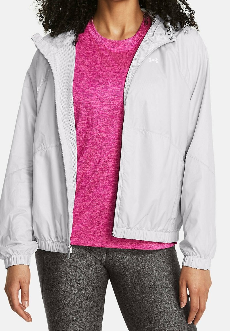 Personne portant un t-shirt de sport rose vif, une veste zippée gris clair et des leggings gris foncé, debout les mains détendues le long du corps.