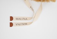 Δύο μπεζ υφασμάτινα λουριά με την επιγραφή "MALINA" σε μαύρο, το καθένα καταλήγοντας σε καφέ δερμάτινο σημείο, σε λευκό φόντο.