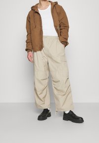 Brun hættejakke, hvid tanktop, beige cargo-bukser med lommer, sorte snørebåndssko, hvide sokker, afslappet pasform, ensfarvede, minimalistisk design.