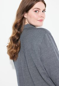 Blazer de patrón de espiga gris con un diseño entallado, cuello de muesca y corte relajado. Presenta pliegues en los hombros y una textura de tela suave.