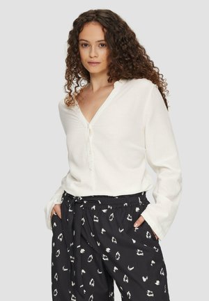 Blouse à manches longues de couleur crème avec un décolleté en V et des détails de boutons. Associée à un pantalon noir avec un motif abstrait blanc.