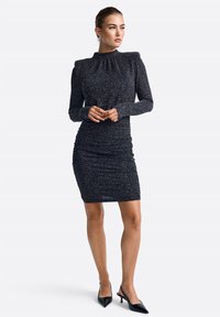 Vestido negro, texturizado, de manga larga con silueta ajustada, escote fruncido y dobladillo a la altura de la rodilla. Combinado con tacones negros de punta.