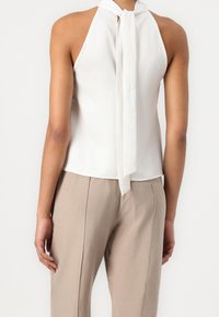 Femme portant un haut sans manches blanc avec un noeud au cou et un pantalon taille haute beige clair, vue de dos.