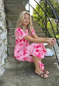 Ung kvinde i lyserød blomsterkjole sidder på udendørs stentrin ved siden af en tekande og en kop, med grønt løv og sort gelænder i nærheden.