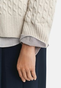 Main détendue sur le côté, portant un pullover en tricot crème avec un motif en côtes par-dessus une chemise rayée et un pantalon bleu foncé, le tout sur fond blanc.