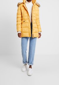 Person wearing a yellow hooded puffer jacket with fur trim, white sweater, cuffed light blue jeans, and white sneakers against a white background.