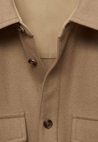 Gros plan sur le devant d'un manteau beige montrant le col, deux boutons et des poches à rabat en tissu doux.