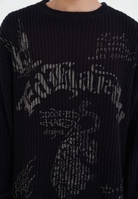 Maglione nero a costine con audaci stampe grafiche in grigio, incluse scritte e disegni astratti. Collo rotondo con maniche lunghe.