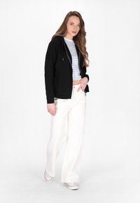 Schwarze Zip-up Kapuzenjacke über einem gestreiften Crop-Top, kombiniert mit weißen weit geschnittenen Hosen und Sneakers aus Canvas. Glatter Stoff und lässiges Design.