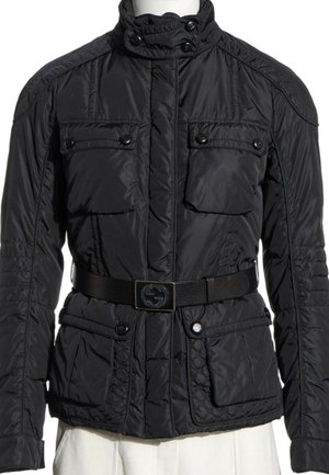Veste matelassée noire avec col montant, quatre poches à rabat boutonnées et ceinture noire avec boucle logo, portée sur un pantalon blanc.