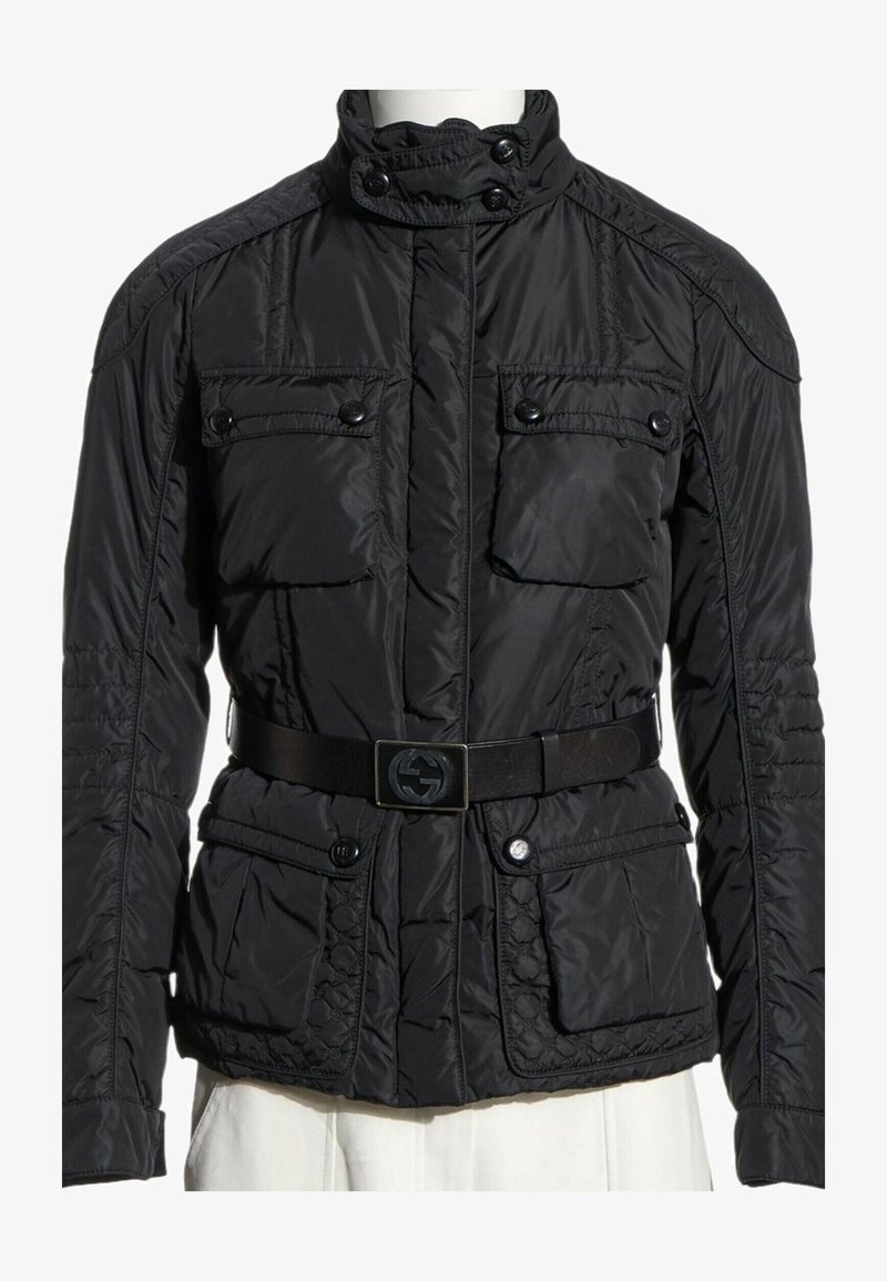 Veste matelassée noire avec col montant, quatre poches à rabat boutonnées et ceinture noire avec boucle logo, portée sur un pantalon blanc.
