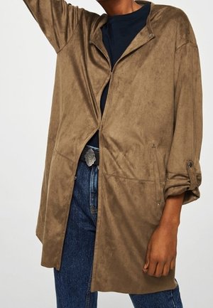 Persona con una chaqueta de ante marrón claro y holgada sobre una camiseta negra y vaqueros azules, con las mangas remangadas y un brazo levantado.