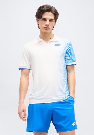 Νέος άνδρας στέκεται φορώντας ένα αθλητικό πόλο με λευκό και μπλε μοτίβο και φωτεινά μπλε σορτς, με χαλαρά χέρια και ουδέτερη έκφραση.