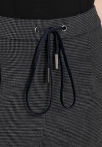 Kalhoty s šedým houndstooth vzorem, se stahovací šňůrkou v pase, navržené s námořnickými kordy a metalickými koncovkami, obsahující boční kapsy a texturovanou tkaninu.