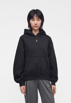Persona che indossa una felpa nera con cappuccio con un logo bianco e pantaloni della tuta grigi con strisce bianche, in piedi davanti a uno sfondo bianco uniforme.