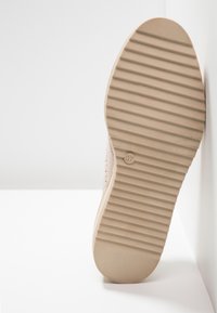 Semelle d'une chaussure beige avec des rainures horizontales et la taille 37, appuyée contre une surface verticale blanche.
