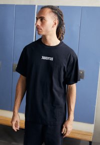 Black cotton t-shirt with short sleeves featuring "JUVENTUS" in bold white lettering on the chest, paired with dark pants in a minimal setting.