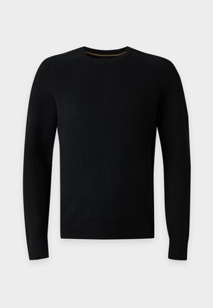 Jeune homme debout portant un pull noir à manches longues, un pantalon marron ample et des chaussures noires, sur un fond blanc uni.
